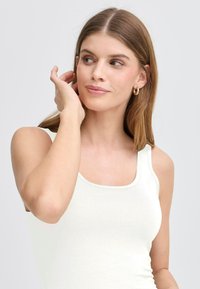 Vit åtsittande linne med fyrkantig halsringning och ribbad struktur. Modellen har långt brunt hår och bär små guldörhängen.