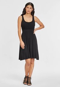 Schwarzes ärmelloses Kleid aus weichem Stoff, mit figurbetontem Oberteil und ausgestelltem Rock. Strukturierter Taillenbund und dezente Raffungen am Ausschnitt.