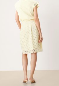Krajková sukně v jemně žluté barvě s květinovými výřezy, vlněným lemem a úzkým pasem. Kombinována s volně střiženým topem světlejší barvy.