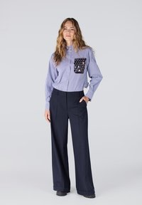 Gestreepte blauwe button-down shirt met een zwarte, pailletten pocket; gecombineerd met wijde, vloeiende marineblauwe broek en zwarte schoenen.