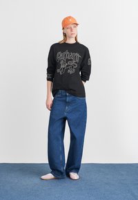Black long-sleeve shirt with white graphic, blue loose-fit jeans, and orange cap. Model wears pink slip-on shoes, standing against a light background.