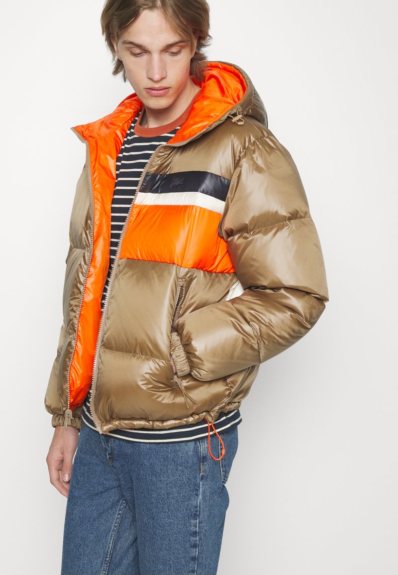 Braune Puffjacke mit orangefarbenen und schwarzen Akzenten, ausgestattet mit Kapuze, Reißverschluss und gestreiften Bündchen. Glatte, glänzende Textur.