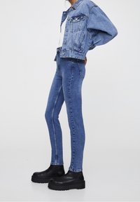 Chaqueta de mezclilla con un corte holgado, de color azul claro, con grandes bolsillos frontales, combinada con jeans skinny de tiro alto en un azul más oscuro y botas de plataforma negras.
