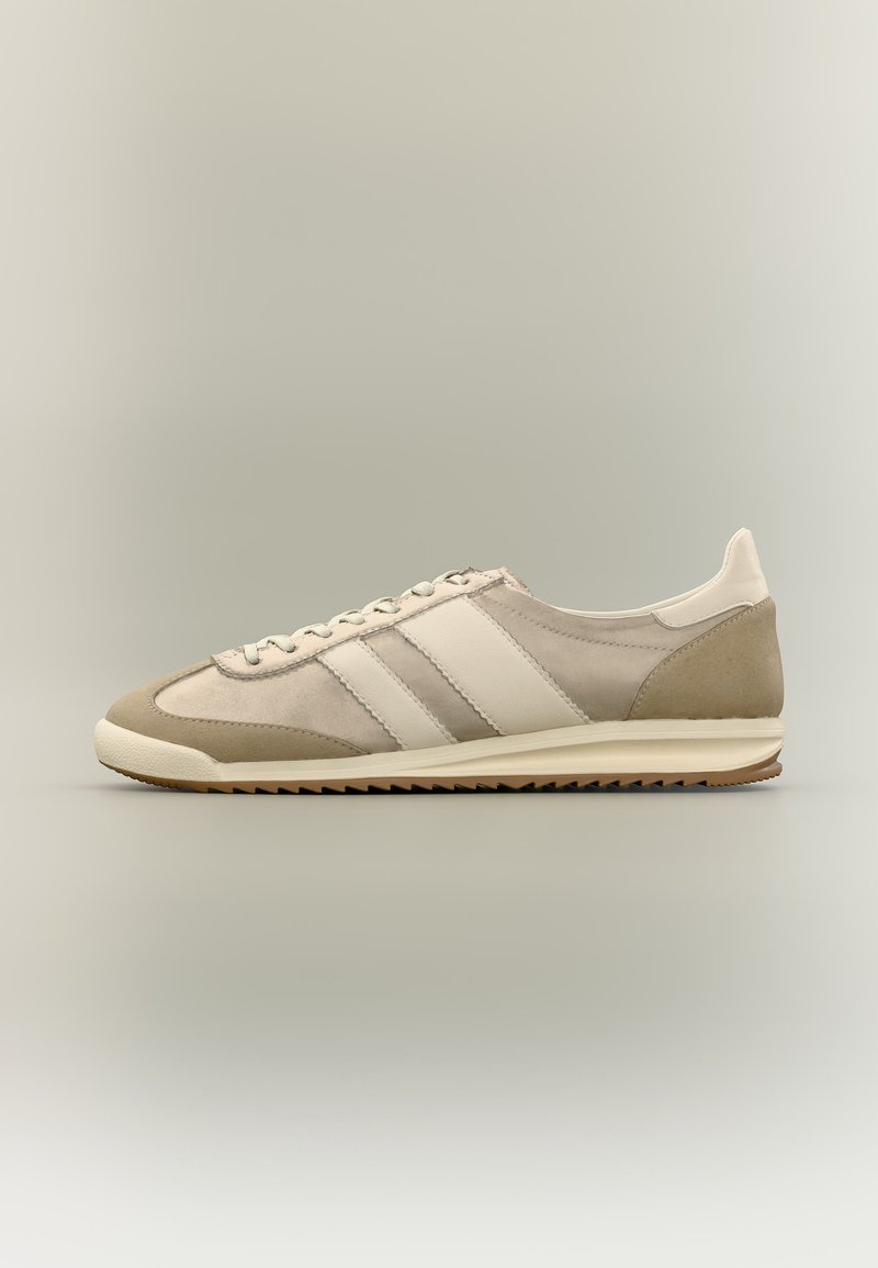 Χαμηλού προφίλ μπεζ και κρεμ αθλητικό παπούτσι με κορδόνια μπροστά, πλαϊνές ρίγες και υφή από καουτσούκ στη σόλα σε ουδέτερο φόντο.