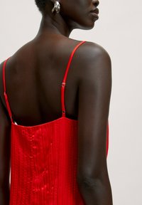 Rode geplooide jurk met dunne verstelbare bandjes en een gladde glans. Dichtbijzicht van de achterkant met detailing, met blootstelling van de huidkleur en schouder.