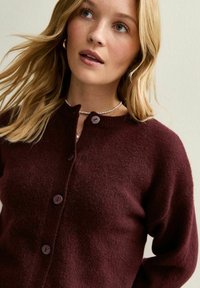 Cardigan en tricot bordeaux avec un col rond, fermeture à quatre boutons et une texture douce. Présente des manches longues et une coupe décontractée.