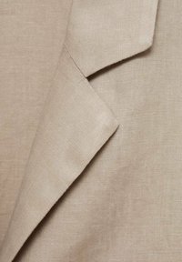 Beige linen fabric showing a folded collar with a subtle texture. The material appears lightweight and breathable with a natural finish.