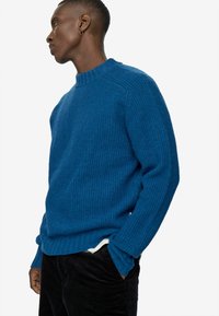 Zils ribotais džemperis ar garām piedurknēm un augstu apkakli, valkāts virs melnajiem kordūrāna biksēm. Audums izskatās teksturēts un biezs.