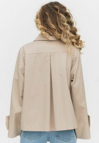 Beżowa kurtka z plisą na plecach, kołnierzem klapowym i szerokimi rękawami. Wykonana z gładkiego materiału, charakteryzująca się czystymi liniami i brakiem widocznych elementów metalowych.
