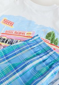 T-shirt avec illustration colorée d'un diner avec des enseignes "Tony's Diner", "Burgers Hot dogs" et "Shakes", associé à un short à taille élastique bleu à carreaux.