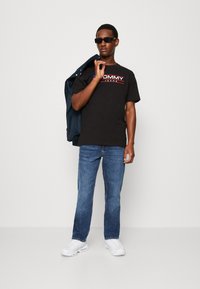 Černé bavlněné tričko s červeným a bílým logem "TOMMY JEANS", doplněné modrými džínami a bílými teniskami. Model drží bundu na rameni.