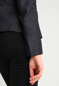 Mörk denimjacka med strukturerad textil, figursydd design och en enda svart knapp på ärmen. Närbild av ärmen och handen.