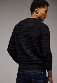 Schwarzer, strukturierter Pullover mit erhöhten horizontalen Streifen, gerippten Bündchen und Saum, getragen von einem Modell, das von hinten zu sehen ist, kombiniert mit blauen Jeans.