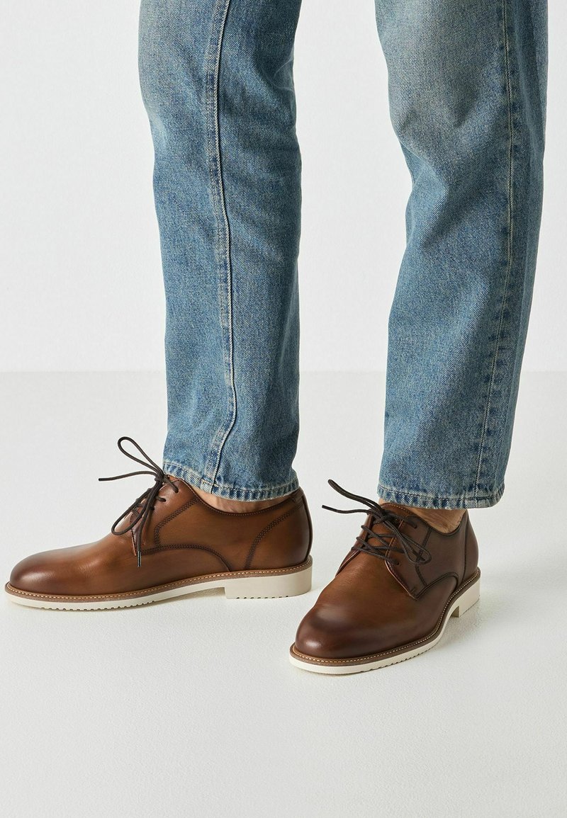 Chaussures à lacets en cuir marron avec une finition lisse, semelle blanche texturée, et accents perforés subtils, portées avec un jean bleu clair.
