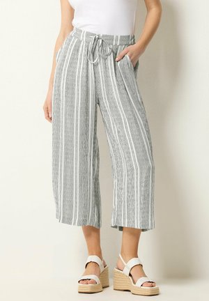 Kvinde iført grå og hvide vertikalt stribede viddebukser med snøre i taljen og hvide kilehælssandaler.