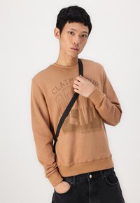 Barna pulóver texturált "DENIM" mintával, kerek nyakkal és bordázott mandzsettákkal. Fekete nadrággal és fekete keresztpánttal párosítva.