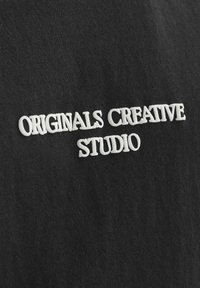 Zwarte stof met witte geborduurde tekst die "ORIGINALS CREATIVE STUDIO" zegt. Soepele textuur met een casual ontwerp.