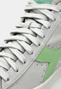 Szürke velúr sneaker, fehér fűzőkkel, texturált felülettel és figyelemfelkeltő zöld logóval. Perforációk emelik ki a dizájnt.