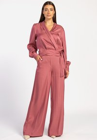 Roze satijnen wikkeltop met lange mouwen, gecombineerd met bijpassende wijde broekspijpen. Heeft een strik in de taille en zakken in de broeken.