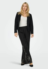 Črni blazer s prilegajočim dizajnom, kombiniran s črnimi širokimi hlačami z belo cvetlično vezenino. Model nosi belo majico brez rokavov.