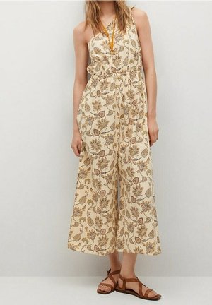 Vrouw draagt een mouwloze crèmekleurige jumpsuit met bloemenprint, gecombineerd met bruine platte sandalen en een amberkleurige hangersketting.