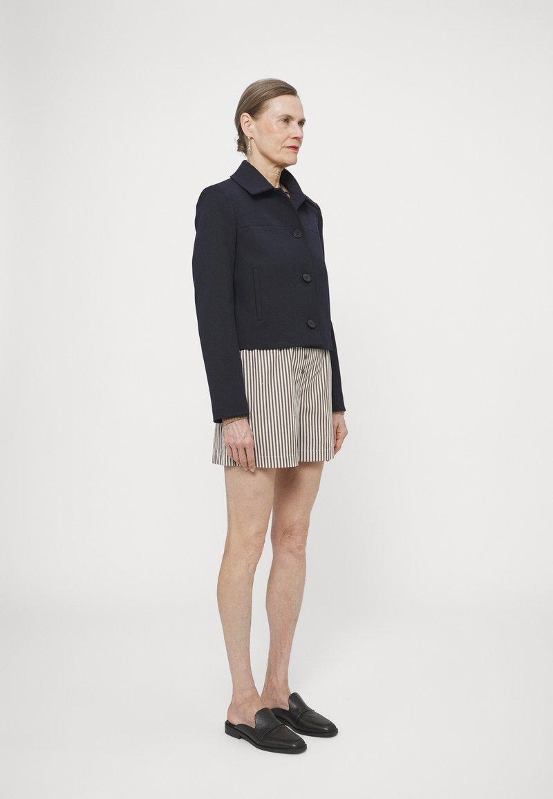Veste cropped navy en tissu texturé, avec un col, des boutons à l'avant et deux poches latérales, associée à un short rayé.
