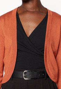 Rostfärgad stickad cardigan med öppen front, lätt textur, bärs över en svart omslagstopp och ett svart bälte med en silverspänne.