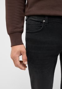 Czarne dżinsy w dopasowanym kroju, z przednią kieszenią, szlufkami na pasek i delikatnym połyskiem. W połączeniu z brązową bluzką z długim rękawem.