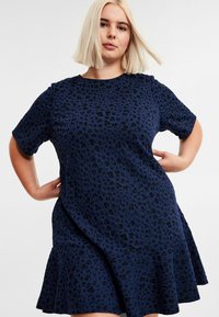 Frau mit blonden Bob-Haaren, die ein navyblaues Kurzarmkleid mit einem schwarzen Leopardenmuster trägt und mit den Händen auf den Hüften vor einem weißen Hintergrund posiert.