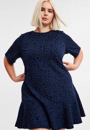 Frau mit blonden Bob-Haaren, die ein navyblaues Kurzarmkleid mit einem schwarzen Leopardenmuster trägt und mit den Händen auf den Hüften vor einem weißen Hintergrund posiert.