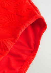 Costume da bagno rosso con motivo testurizzato di conchiglie, caratterizzato da un rivestimento interno liscio e bordi elasticizzati. Vista ravvicinata del bordo della vita.