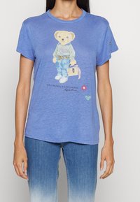 Modré bavlněné tričko s grafikou medvěda oblečeného v svetru a džínech. Na spodní části jsou květinové a srdcové akcenty. Měkká textura.