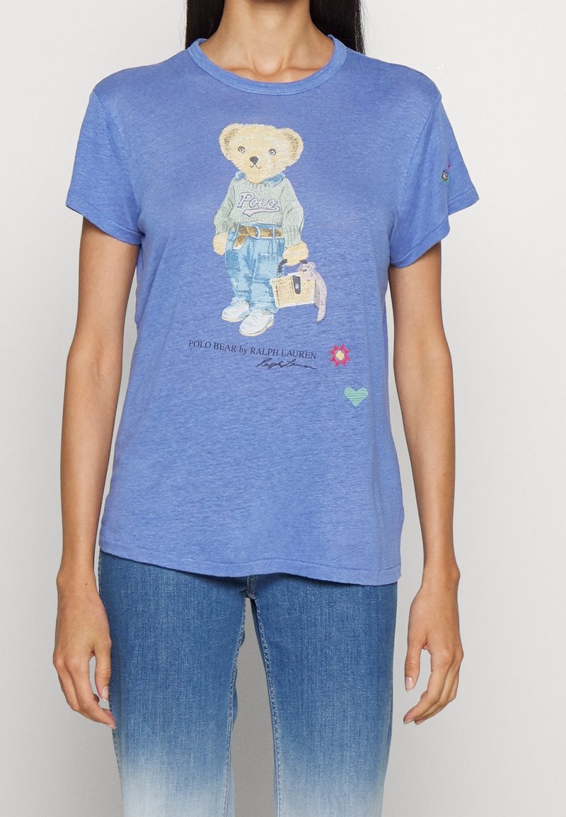 Modré bavlněné tričko s grafikou medvěda oblečeného v svetru a džínech. Na spodní části jsou květinové a srdcové akcenty. Měkká textura.
