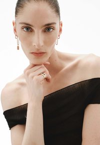 Top negro sin hombros con textura acanalada; la modelo lleva pendientes de aro dorados y un anillo dorado con acentos verdes; fondo neutro.