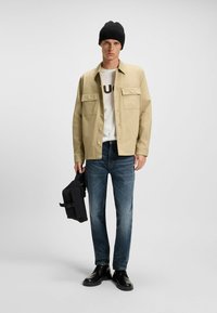 Beige button-up jacket, white T-shirt with dark print, dark jeans, black beanie, holding a small black bag, black boots. Plain background.