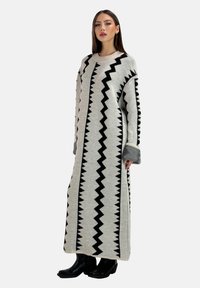 Langes, graues Strickkleid mit schwarzem Zickzackmuster. Es hat einen runden Ausschnitt und weite Ärmel mit kontrastierenden grauen Bündchen. Kombiniert mit schwarzen Stiefeln.