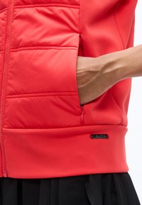 Χέρι που έχει εισαχθεί στην τσέπη με φερμουάρ ενός κόκκινου μπουφάν puffer, με μια μαύρη φούστα με πιέτες να φαίνεται από κάτω.