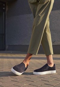 Sapatos de camurça pretos, tipo slip-on, com solas de borracha brancas, combinados com calças cropped em verde-oliva claro. O pavimento apresenta um padrão texturizado.