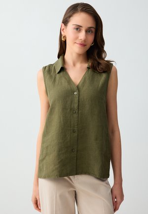 Donna che indossa una camicia verde oliva senza maniche con bottoni e colletto e pantaloni beige, in piedi davanti a uno sfondo chiaro uniforme.