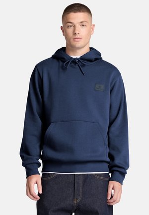 Joven con sudadera con capucha azul marino con bolsillo delantero y vaqueros oscuros, de pie frente a un fondo liso y claro.