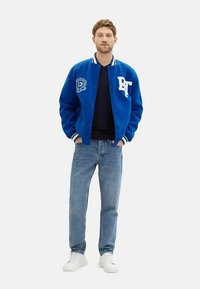 Jachetă varsity albastră cu accente și logo alb, asortată cu un tricou negru, blugi albaștri deschis și adidași albi. Stil sportiv formal.