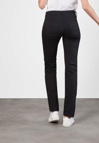 Persoon die strakke zwarte jeans en witte sneakers draagt, staand op een grijze vloer tegen een effen witte achtergrond.