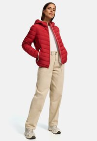 Red padded jacket with a hood, featuring quilted texture and white zipper. Paired with beige pants and light-colored sneakers.