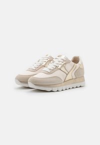 Beige sneakers med en texturerad tyg- och mockakombination. Har guldaccent, vita snörningar och en dämpad sula med ett vågmönster.