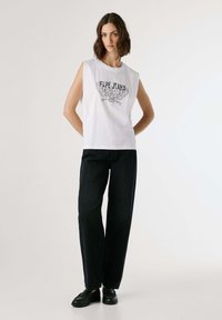 Donna con capelli corti castani che indossa un top senza maniche bianco di Pepe Jeans, pantaloni neri a gamba larga e mocassini neri, su uno sfondo chiaro e semplice.