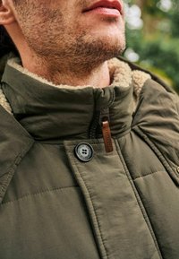 Jachetă matlasată de culoare verde măsliniu, cu guler înalt, căptușeală din fleecă, detalii cu nasturi și un accent din piele maro pe gluga fermoarului.