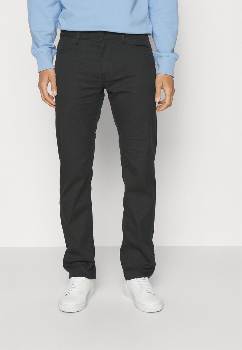 Pantalon noir en tissu extensible, coupe ajustée. Doté de poches avant et d'une silhouette élégante, porté avec des baskets blanches et un sweat-shirt bleu.