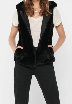 Femme portant un gilet sans manches noir et duveteux par-dessus un t-shirt blanc à manches courtes avec col en V et un pantalon noir, les mains dans les poches du gilet.