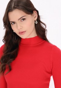 Maglione a collo alto rosso a coste con maniche lunghe. Tessuto liscio e elastico. Modella con orecchini a cerchio bianchi. Texture morbida, design aderente.