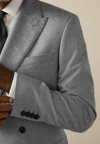 Grau strukturierte Blazer mit Reverskragen, einer einzelnen Brusttasche und vier schwarzen Knöpfen an den Manschetten. Über einem weißen Hemd und einer Krawatte getragen.